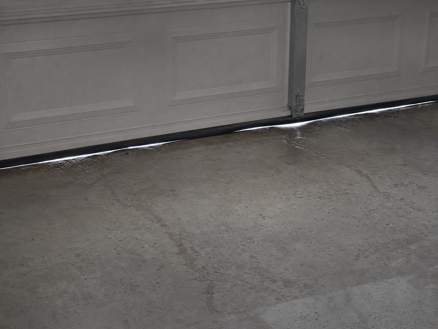 A wet track of water along the base of a closed sectional garage door on a concrete floor